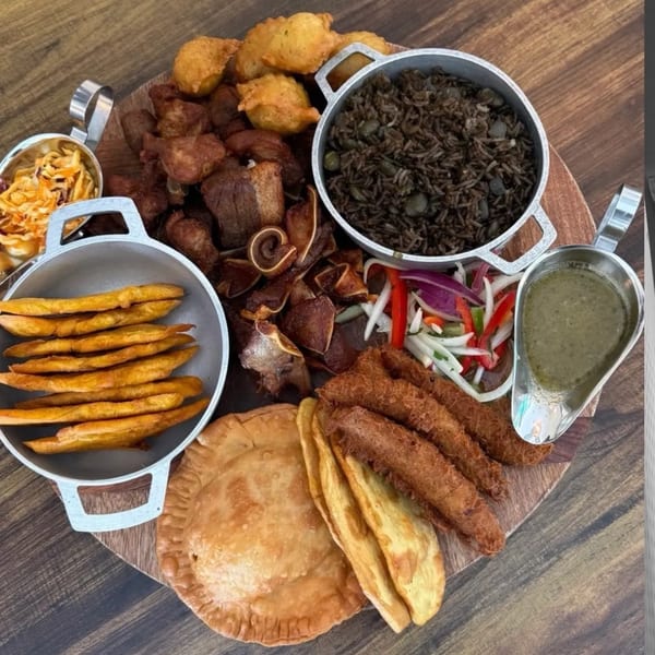 A large Haitian platter with fried pork, black rice, plantains, fritters, and pikliz on a wooden board.
