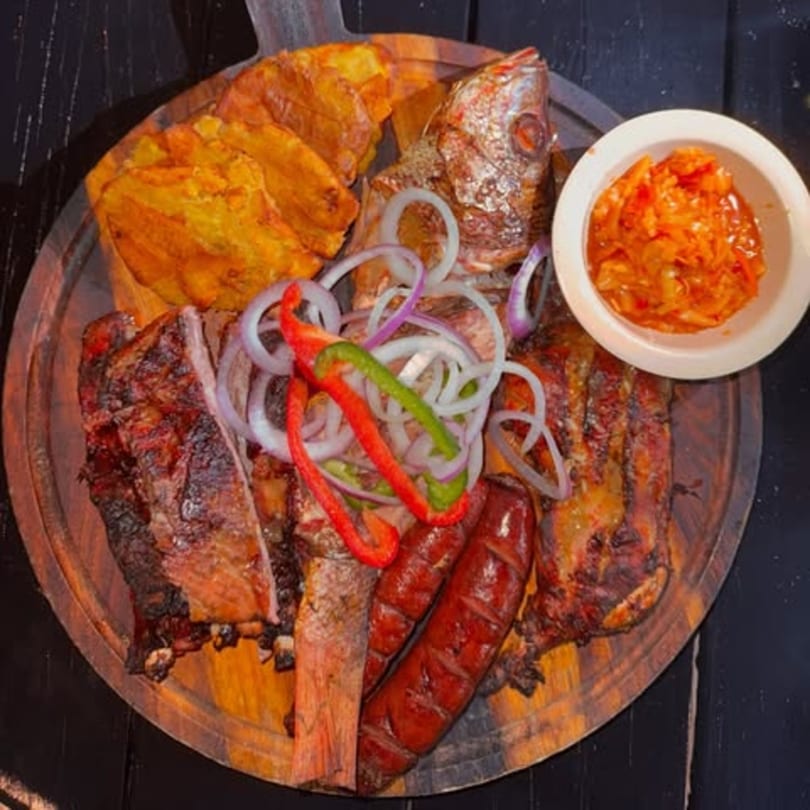 A Haitian griot platter with fried fish, ribs, sausage, plantains, and pikliz on a wooden board.