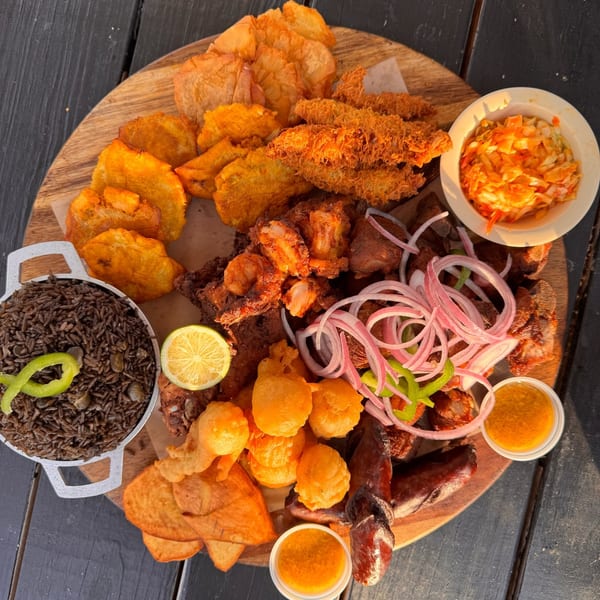 A large Haitian platter with fried plantains, rice, meat, and pikliz on a wooden board.