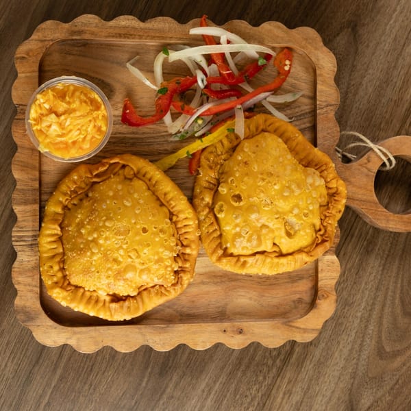 Two golden Haitian patties with a side of pikliz and a dipping sauce on a wooden tray.