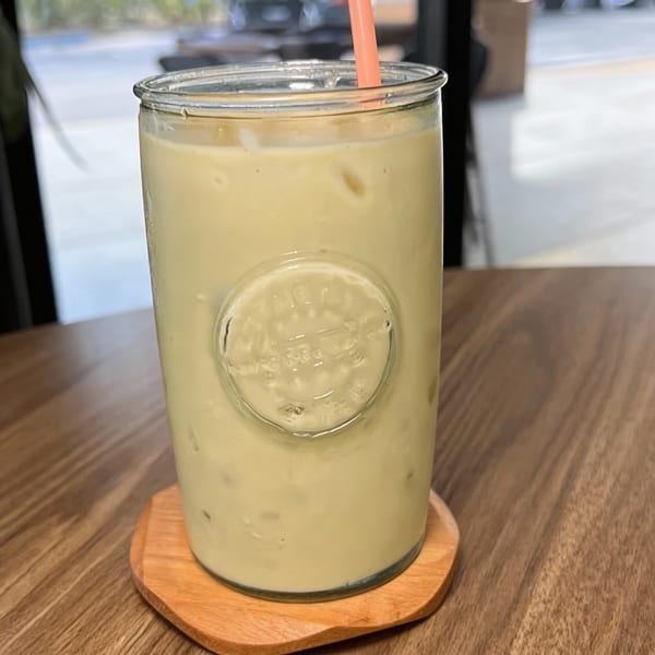 A glass of light green Haitian drink with ice and a pink straw on a wooden coaster.