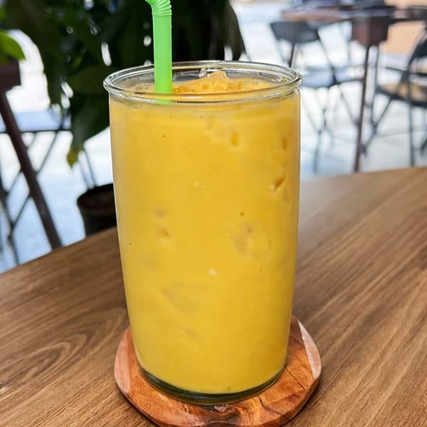 A vibrant yellow mango smoothie with a green straw, on a wooden table at Sak Pase Kitchen.