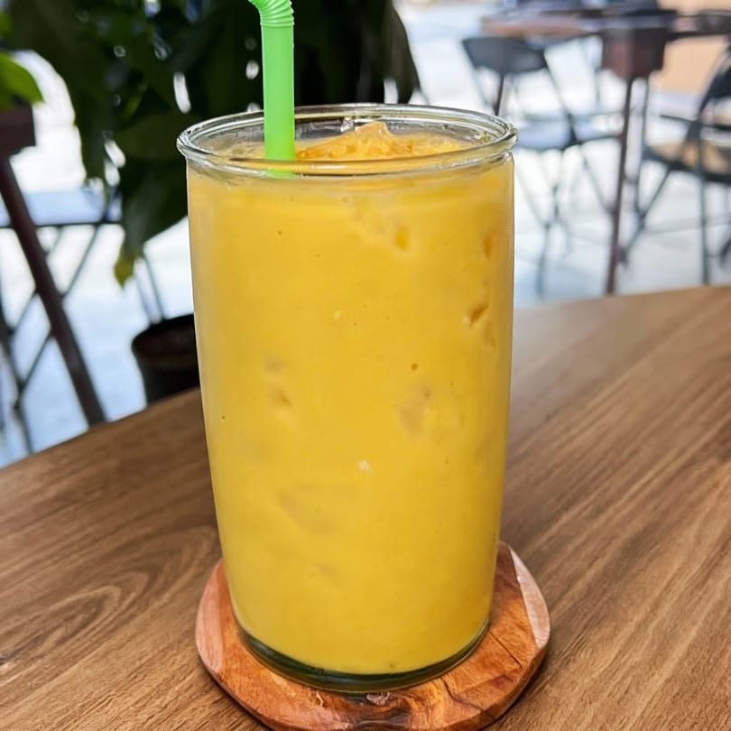 A vibrant yellow mango smoothie with a green straw, on a wooden table at Sak Pase Kitchen.