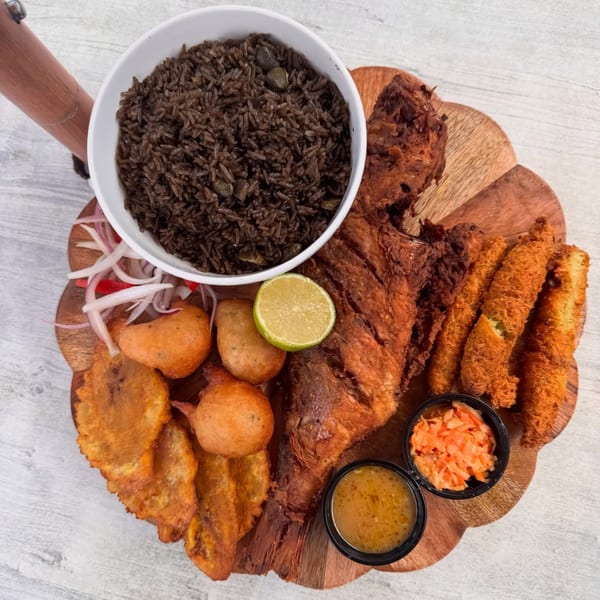 Haitian meal with fried fish, black rice, plantains, fritters, and sauces on a wooden board.
