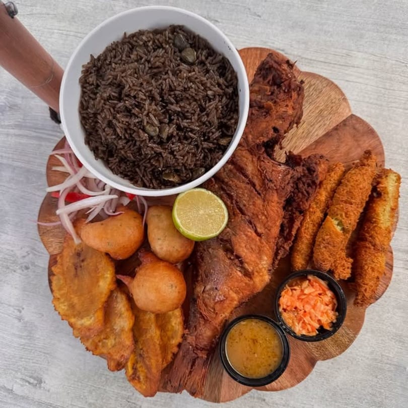 Haitian fried fish platter with black rice, plantains, fritters, pikliz, and dipping sauces on a wooden board.
