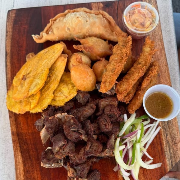 Haitian griot, fried plantains, accra, and pate kode served on a wooden board with sauces.