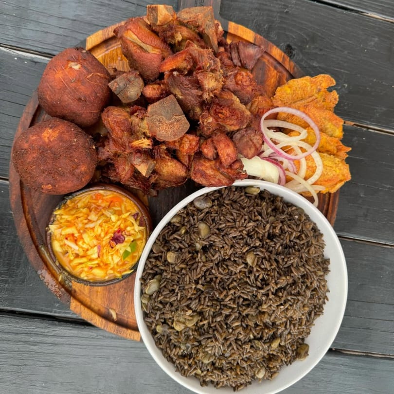 Haitian griot with fried plantains, pikliz, and black mushroom rice on a wooden platter.