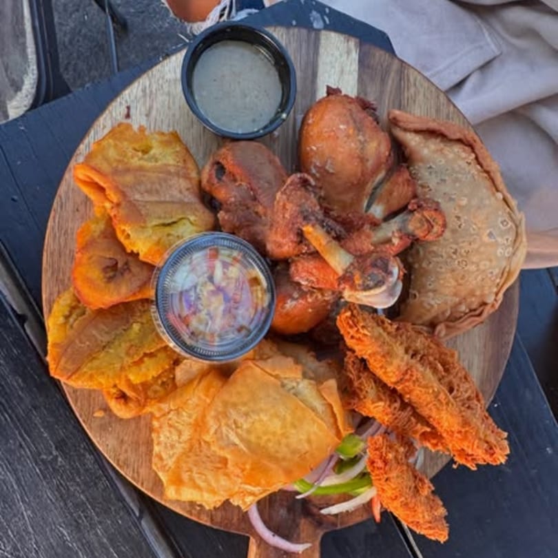 A wooden platter with Haitian fried chicken, plantains, fried pork, and sauces on a dark table.