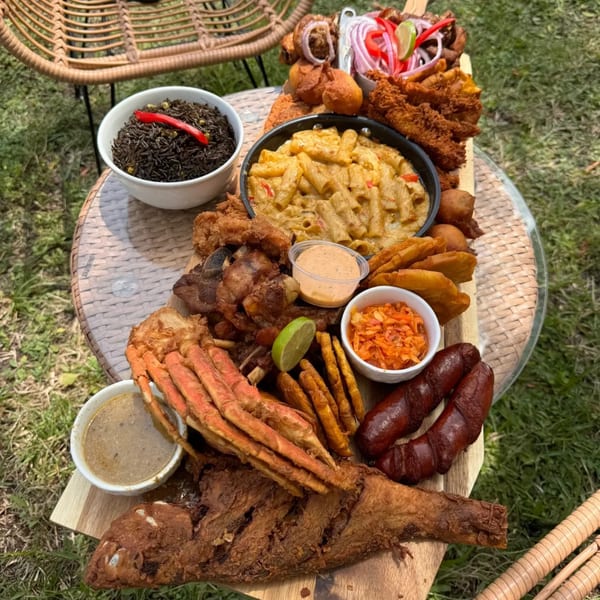 A large platter of Haitian food, including fried fish, crab, pasta, rice, and plantains, on an outdoor table.