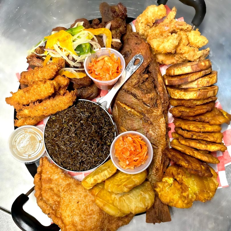 A large platter of Haitian food, including fried fish, rice, plantains, and various fried meats.